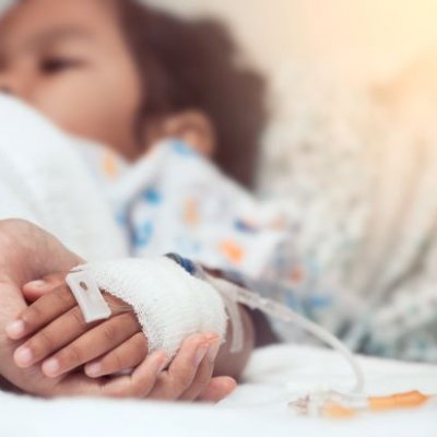 An adult hand holds a bandaged child's hand in the foreground, the child lays in a hospital bed.